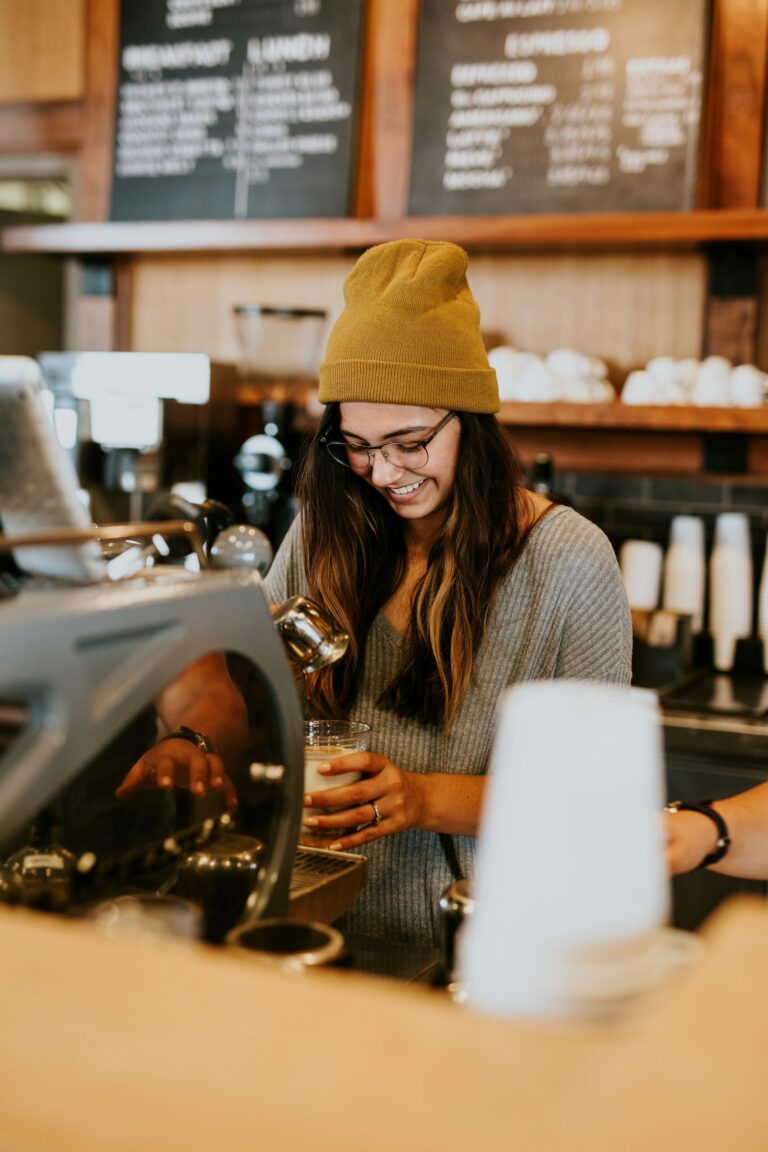 Woman behind a bar pouring a drink. She is wearing a woolly hat.