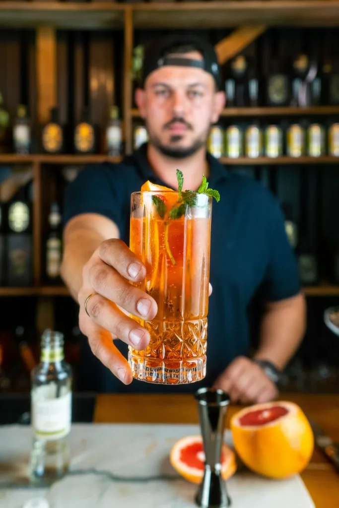 A Barman holding a tall drink out in front of him.